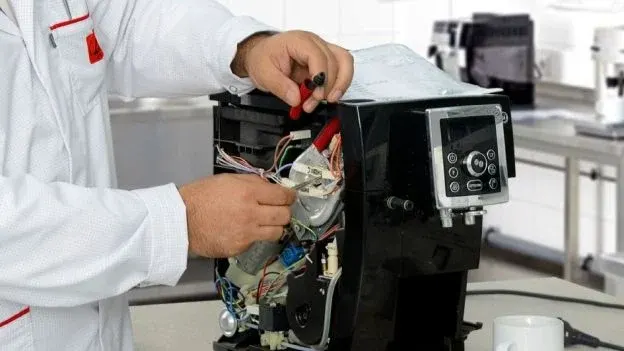 Osoba w białym fartuchu naprawia czarny ekspres do kawy, używając czerwonych szczypiec. W tle widoczne inne urządzenia kuchenne.