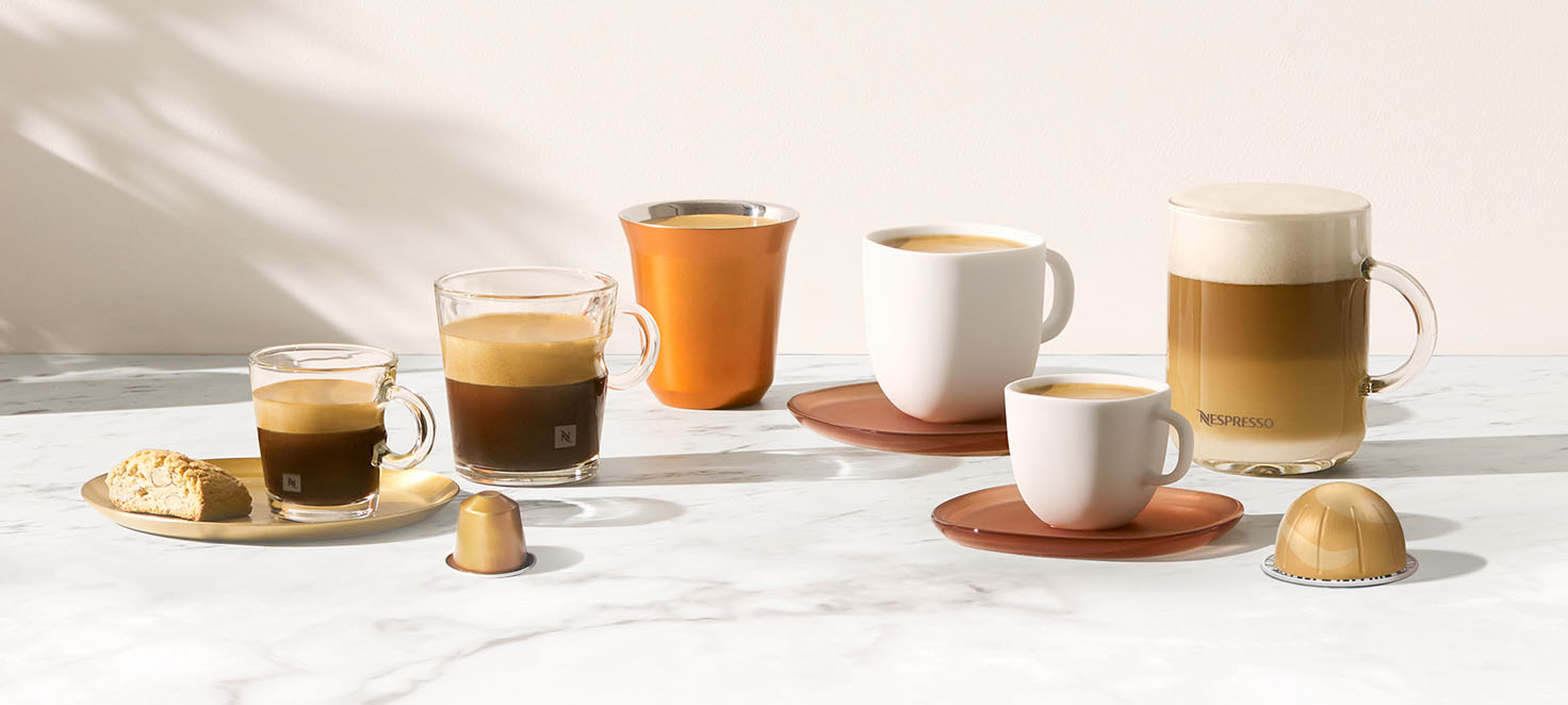 Różne kawy w szklankach i filiżankach na marmurowym blacie z kapsułkami kawy i ciastkiem na złotej tacy.