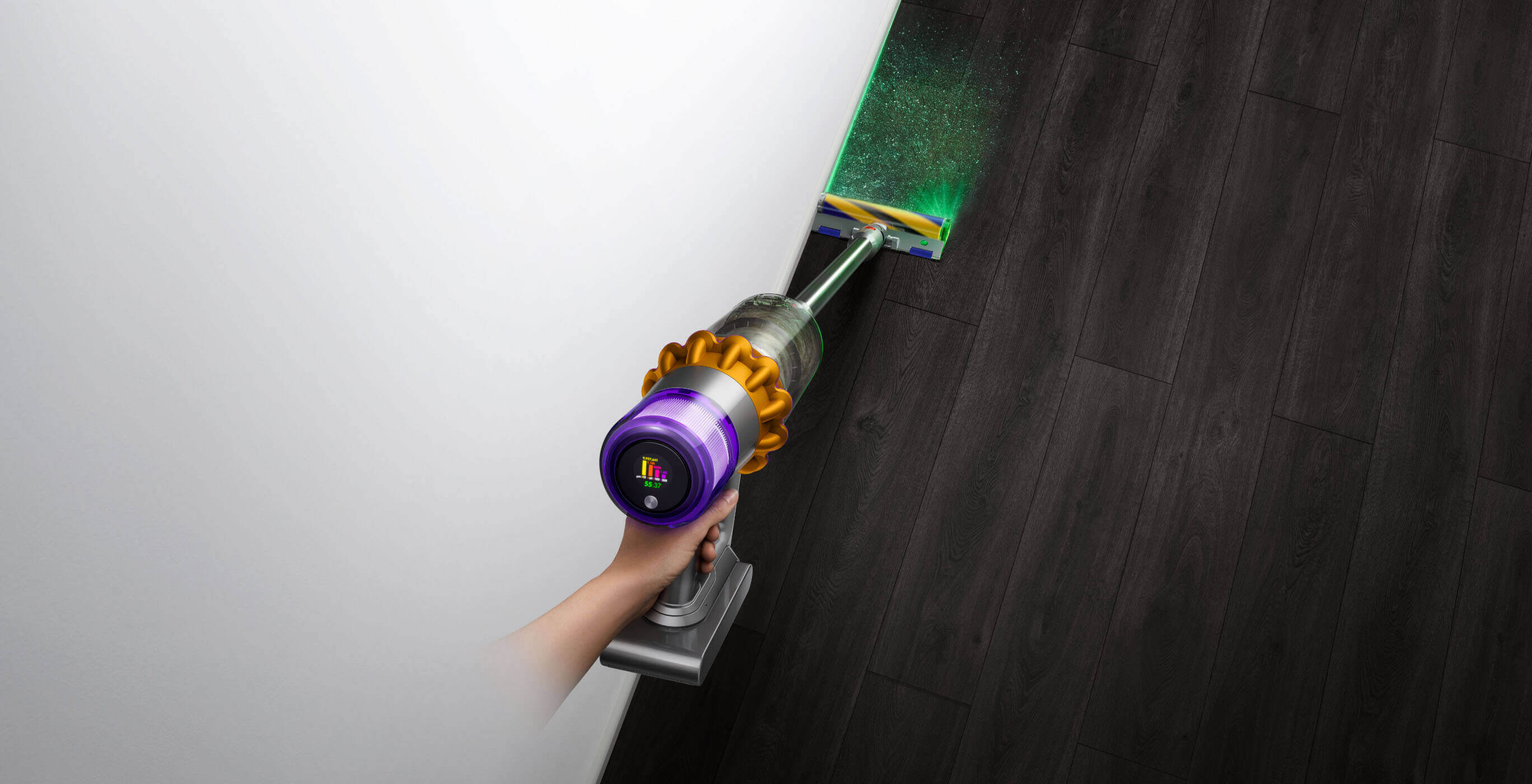Osoba odkurza podłogę odkurzaczem bezprzewodowym Dyson z podświetleniem, przy białej ścianie.