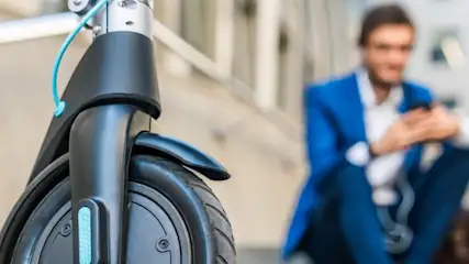 Na pierwszym planie hulajnoga elektryczna, w tle siedzący mężczyzna w niebieskim garniturze, trzymający telefon.