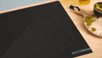 Czarna płyta indukcyjna z czterema polami grzejnymi na drewnianym blacie, obok deska do krojenia z jedzeniem.