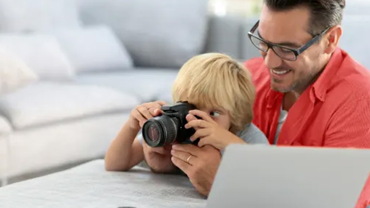 Ojciec w okularach patrzy, jak dziecko robi zdjęcie aparatem fotograficznym; obok biały laptop i sofa w tle.