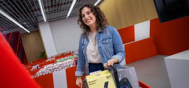 Kobieta niesie torbę z zakupami w sklepie z czerwonymi i białymi ścianami. Widoczna sufitowa konstrukcja i oświetlenie.