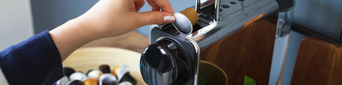 Ręka wkłada kapsułkę kawy do srebrno-czarnego ekspresu. W tle widoczna misa z kapsułkami i fragment drewnianego blatu.