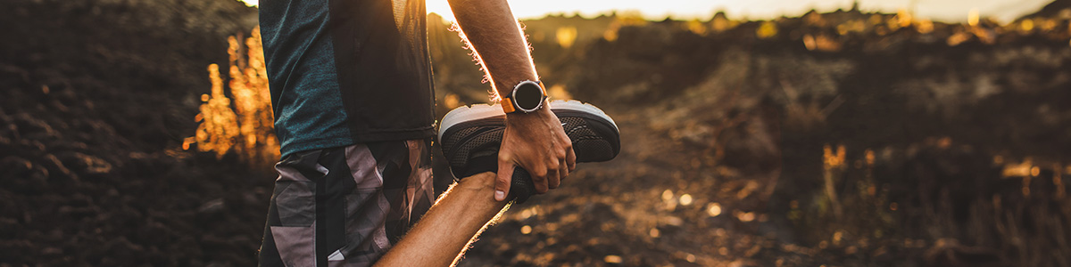 Osoba w sportowym stroju rozciąga nogę, trzymając buta. W tle pagórkowaty krajobraz i zachodzące słońce.