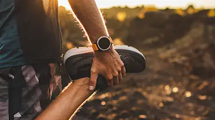 Osoba w sportowym stroju rozciąga nogę, trzymając buta. W tle pagórkowaty krajobraz i zachodzące słońce.