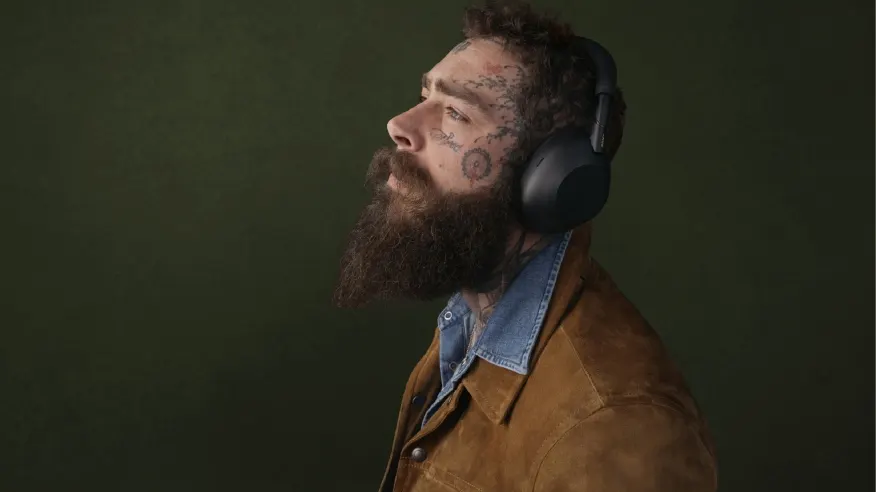Mężczyzna z brodą i tatuażami na twarzy ma na głowie czarne słuchawki. Ubrany jest w brązową kurtkę i dżinsową koszulę. Tło jest ciemnozielone.