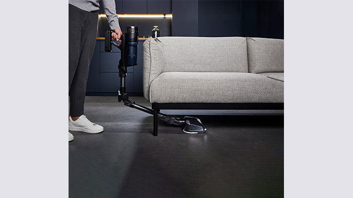 Osoba odkurza podłogę odkurzaczem akumulatorowym przy szarej sofie na tle ciemnej ściany i podłogi.