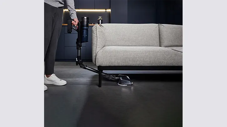 Osoba odkurza podłogę odkurzaczem akumulatorowym przy szarej sofie na tle ciemnej ściany i podłogi.