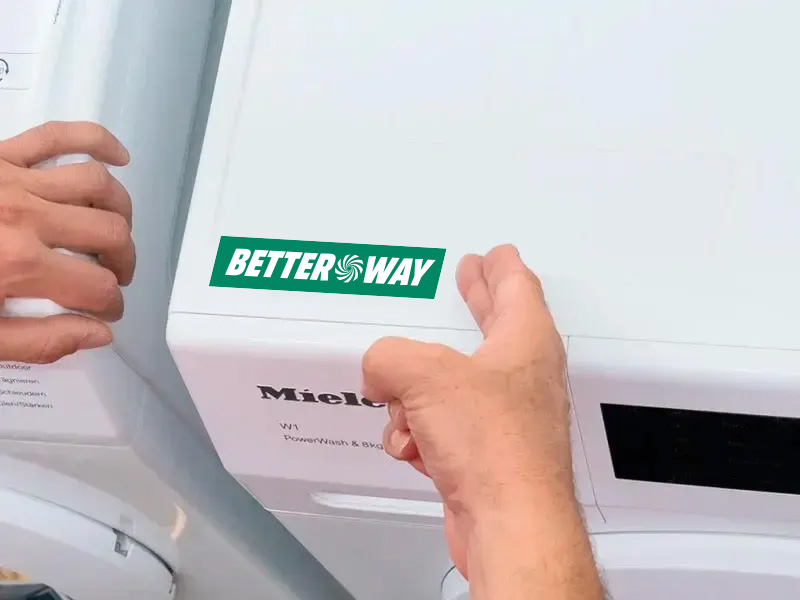 Dłonie dotykają białej pralki Miele z zielonym logo Better Way. Widoczny czarny panel sterowania.