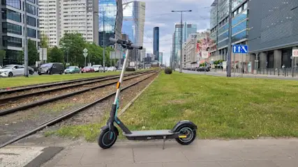 Hulajnoga stoi obok torów tramwajowych na chodniku. W tle budynki miejskie i ulica.