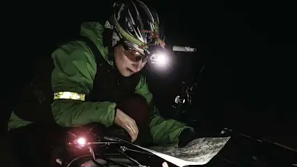 Osoba w kasku i okularach czyta mapę przy rowerze w nocy, oświetlona lampą na głowie i rowerze. Kurtka zielona z odblaskiem.