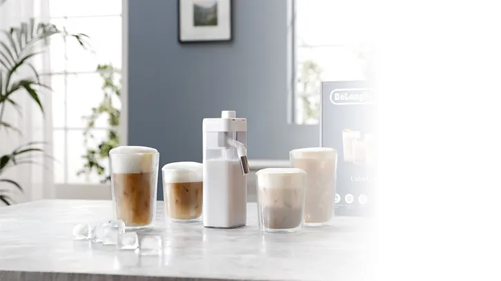 Na stole stoją szklanki z kawą, biały spieniacz do mleka i kostki lodu. W tle okno, roślina i obraz.