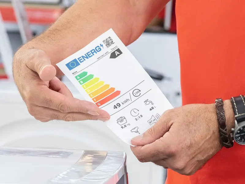 Mężczyzna trzyma etykietę energetyczną A z kolorowymi paskami i danymi o zużyciu energii elektrycznej.