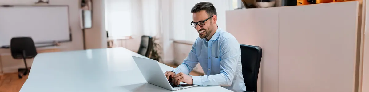 Mężczyzna w okularach siedzi przy białym stole i używa laptopa w biurze. Za nim szafka i krzesła.