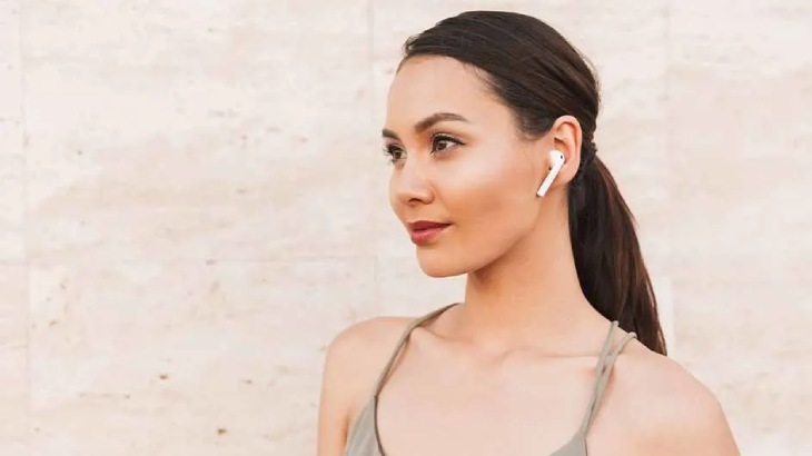 Kobieta z włosami związanymi w kucyk, ma w uchu białą słuchawkę. Ubrana jest w szary top na ramiączkach, tło jest beżowe.