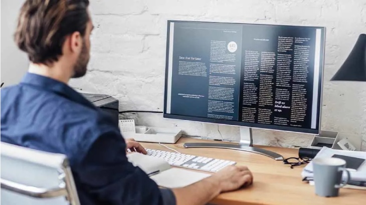 Osoba w niebieskiej koszuli pisze na klawiaturze przed monitorem z tekstem. Na biurku kubek i okulary.