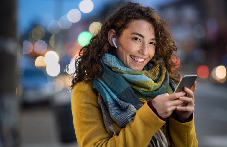 Uśmiechnięta kobieta w słuchawkach trzyma telefon na tle ulicy z rozmytymi światłami. Ma żółtą kurtkę i szalik w odcieniach niebieskiego.