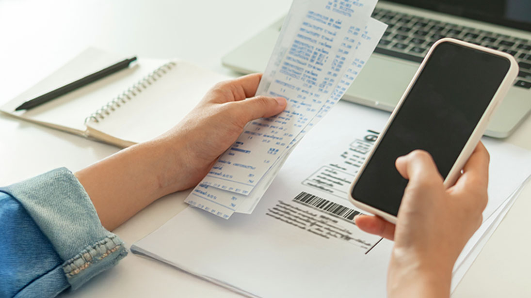 Na stole leżą dokumenty, notatnik, długopis, laptop i telefon. Osoba trzyma paragony i dokument ze skanerem kodu kreskowego.