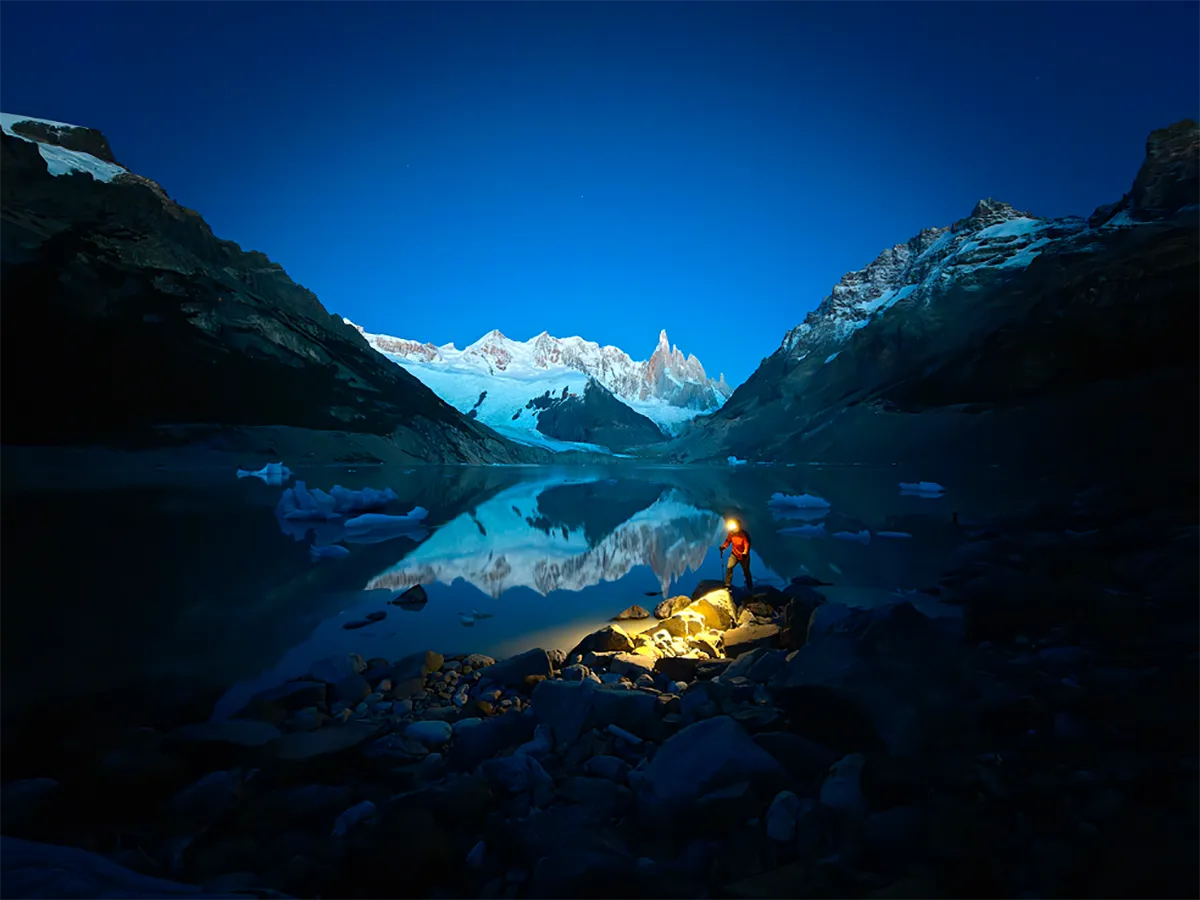 Zmrok w górach: ośnieżone szczyty odbijają się w spokojnym, lodowcowym jeziorze; na pierwszym planie osoba w czerwonej kurtce oświetla skały latarką, na wodzie unoszą się kry.