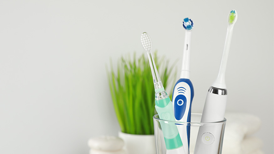 Trzy szczoteeczki elektryczne w szklance: dwie soniczne (biała premium i mniejsza transparentna) i jedna rotacyjna. W tle roślina doniczkowa.