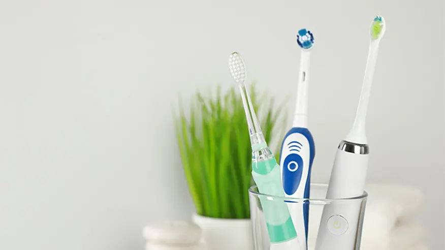 Trzy szczoteeczki elektryczne w szklance: dwie soniczne (biała premium i mniejsza transparentna) i jedna rotacyjna. W tle roślina doniczkowa.
