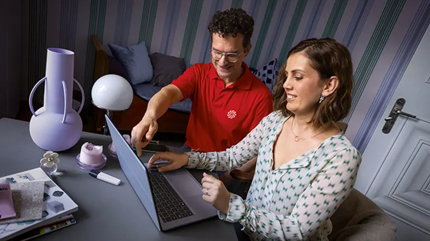 Dwie osoby patrzą na laptopa na biurku z wazonem, lampą i książkami. Mężczyzna w czerwonej koszulce wskazuje na ekran.