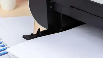 Drukarka drukuje dokument z niebieskimi wykresami słupkowymi. Obok leży lupa z żółtą ramką.