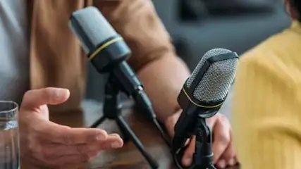 Dwie osoby rozmawiają przy mikrofonach. Na pierwszym planie szklanka wody, mężczyzna gestykuluje ręką. Kobieta w żółtej koszuli.