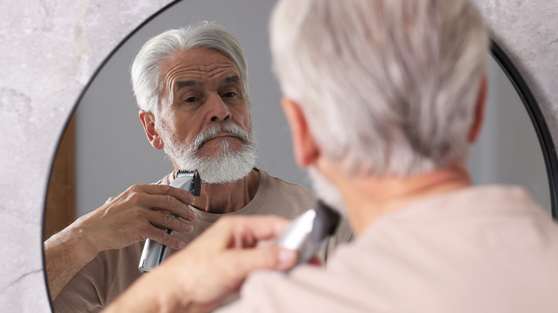 Skupiony, dojrzały i elegancki mężczyzna z bujnym siwym zarostem przycina brodę maszynką elektryczną przed lustrem.