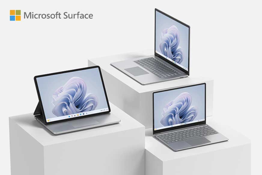 Trzy urządzenia Microsoft Surface na białych podestach. Jeden tablet i dwa laptopy z ekranami skierowanymi do przodu.