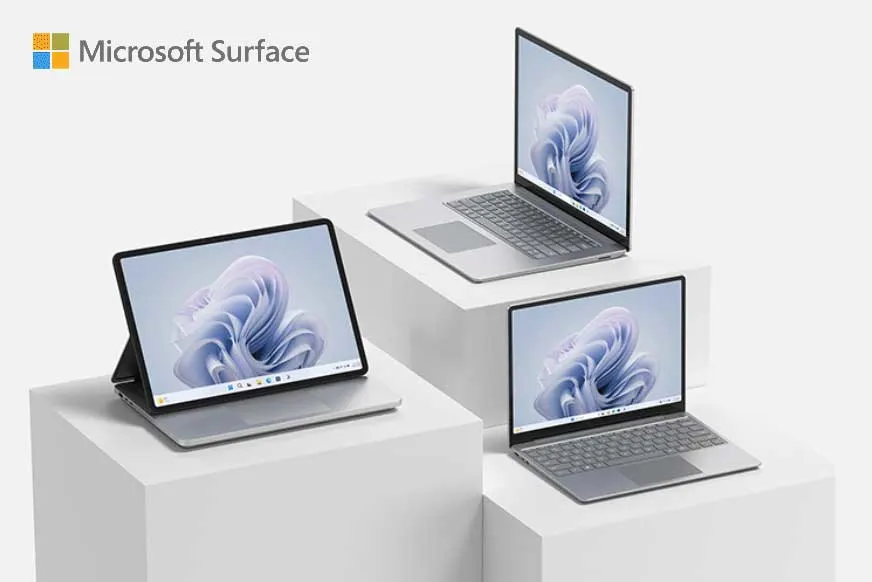 Trzy urządzenia Microsoft Surface na białych podestach. Jeden tablet i dwa laptopy z ekranami skierowanymi do przodu.