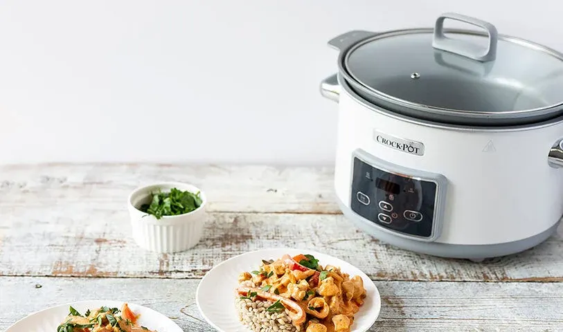 Na drewnianym stole stoi biały, okrągły garnek Crock-Pot z przezroczystą pokrywą, obok miska z zielenią i talerz z potrawą.