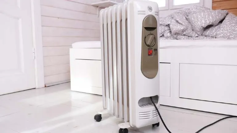 Biały grzejnik olejowy w jasnej sypialni domku na działce z drewnianymi ścianami.