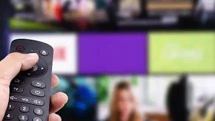 Dłoń trzyma czarny pilot do telewizora. W tle ekran z różnokolorowymi kaflami aplikacji wideo.