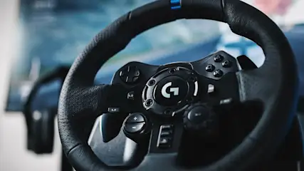 Czarna kierownica do gier z przyciskami i logo "G" na tle niebieskiego ekranu.