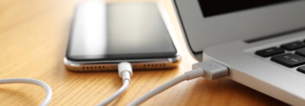 Telefon i laptop podłączone białym kablem do ładowania na drewnianym blacie.