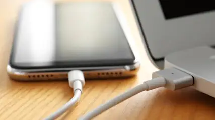 Telefon i laptop podłączone białym kablem do ładowania na drewnianym blacie.