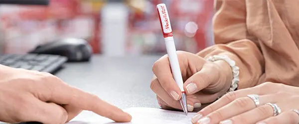 Osoba podpisuje dokument długopisem z czerwonym napisem. Druga osoba wskazuje palcem na dokument. W tle klawiatura i myszka.