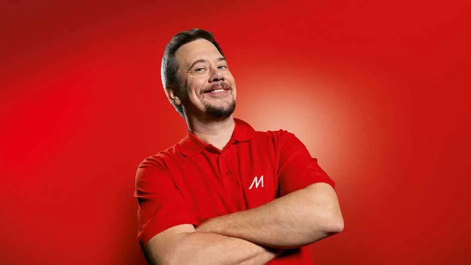 Mężczyzna w czerwonej koszulce polo z literą "M" uśmiecha się, trzymając skrzyżowane ręce na tle czerwonego tła.