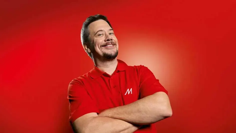 Mężczyzna w czerwonej koszulce polo z literą "M" uśmiecha się, trzymając skrzyżowane ręce na tle czerwonego tła.