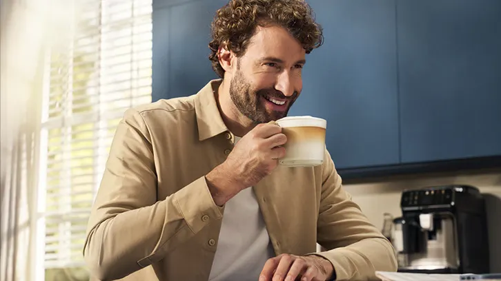 Mężczyzna w beżowej koszuli pije kawę z dużego kubka. W tle okno z żaluzjami i ekspres do kawy.
