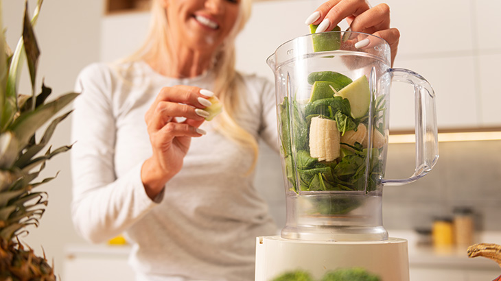 Dojrzała, uśmiechnięta i zadbana kobieta przygotowuje z łatwością zdrowy, zielony koktajl w blenderze, dodając świeże owoce.