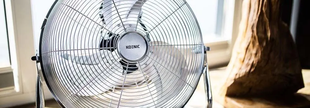 Metalowy wentylator stoi na stole obok okna, a obok niego drewniana rzeźba.