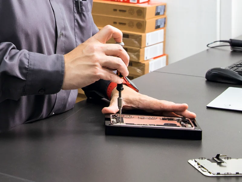 Osoba w szarej koszuli naprawia telefon śrubokrętem na czarnym stole. W tle kartony i akcesoria komputerowe.