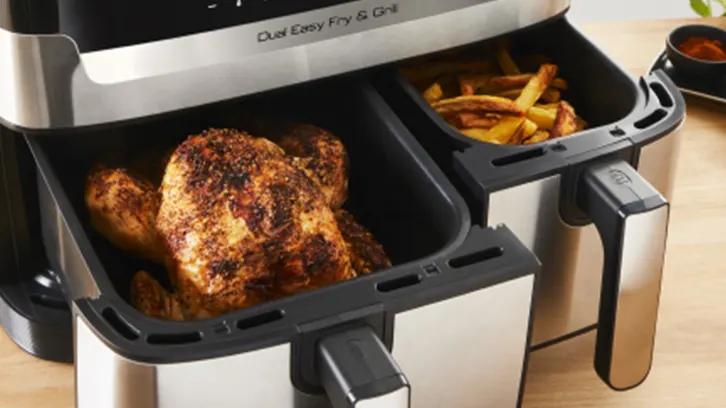 Frytkownica z kurczakiem i frytkami w koszykach. Urządzenie ma srebrną obudowę i czarne uchwyty. Miska z przyprawami w tle.