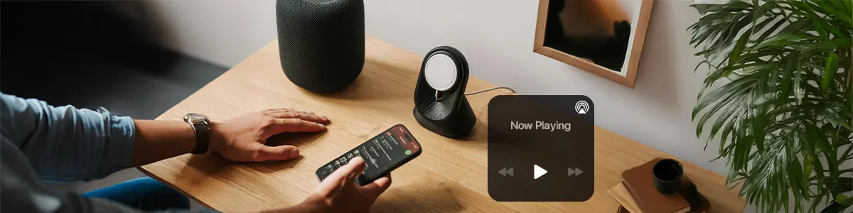 Osoba trzyma jedną rękę na biurku, w drugiej ma iPhone'a. Obok znaczek AirPlay i jakiś głośnik bezprzewodowy