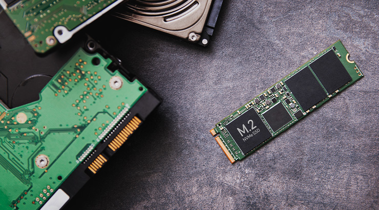 Na szarym tle leżą elementy komputerowe: dysk twardy, zielona płytka drukowana i dysk SSD M.2.