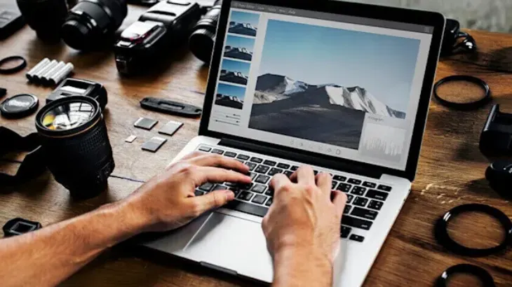 Osoba pisze na laptopie. Na ekranie widać zdjęcie gór. Obok leżą akcesoria fotograficzne: obiektywy i karty pamięci.