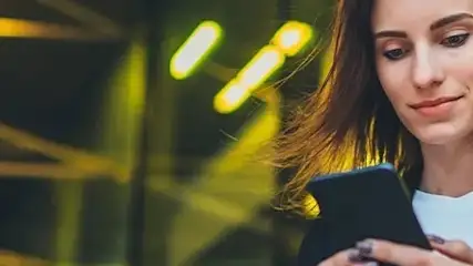 Kobieta w słuchawkach patrzy na telefon na tle miejskiej architektury z żółtymi światłami.